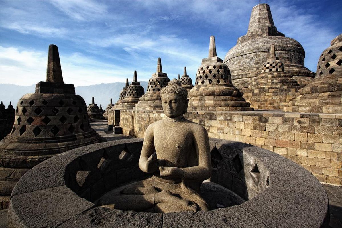 Panorama Candi Borobudur di pagi hari, menunjukkan stupa-stupa Buddha yang megah dengan latar belakang pegunungan hijau dan langit berawan.